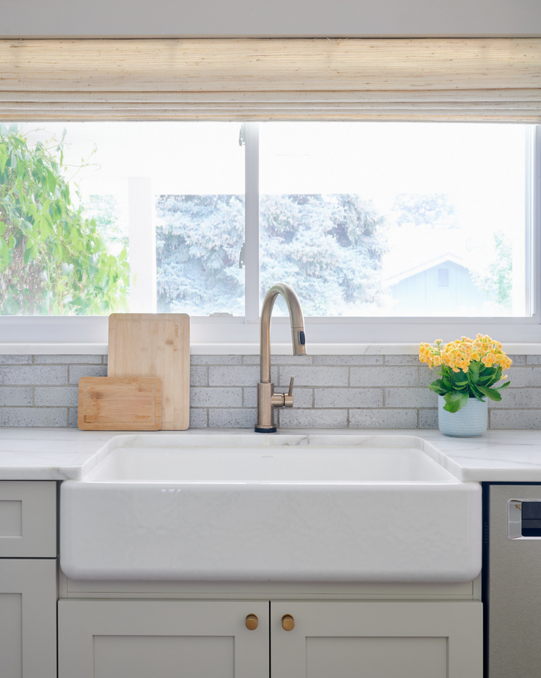 <img src="whitefarmhousesinkunderwindowwithnaturalwovenblinds.png" alt="White farmhouse sink under window with natural woven blinds, light greige cabinets, brass hardware, cutting boards, and flowers">